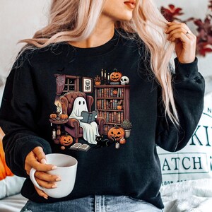 May include: Black sweatshirt featuring a Halloween-themed graphic. The design shows a ghost reading in a cozy armchair, surrounded by pumpkins, a black cat, and a bookshelf. The scene is set in a room with a window and candles.