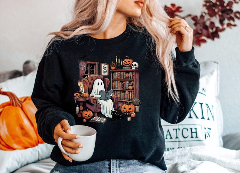 May include: Black sweatshirt featuring a Halloween-themed graphic. The design shows a ghost reading in a cozy armchair, surrounded by pumpkins, a black cat, and a bookshelf. The scene is set in a room with a window and candles.
