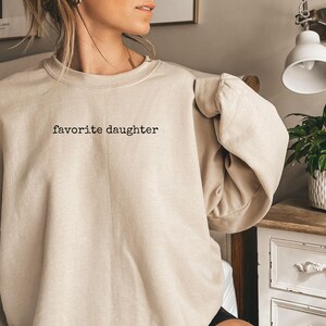 Może przedstawiać: Beżowa bluza z okrągłym dekoltem z czarnym napisem "favorite daughter". Bluza ma luźny kr&oacute;j i długie rękawy. Zdjęcie zrobione w pomieszczeniu.