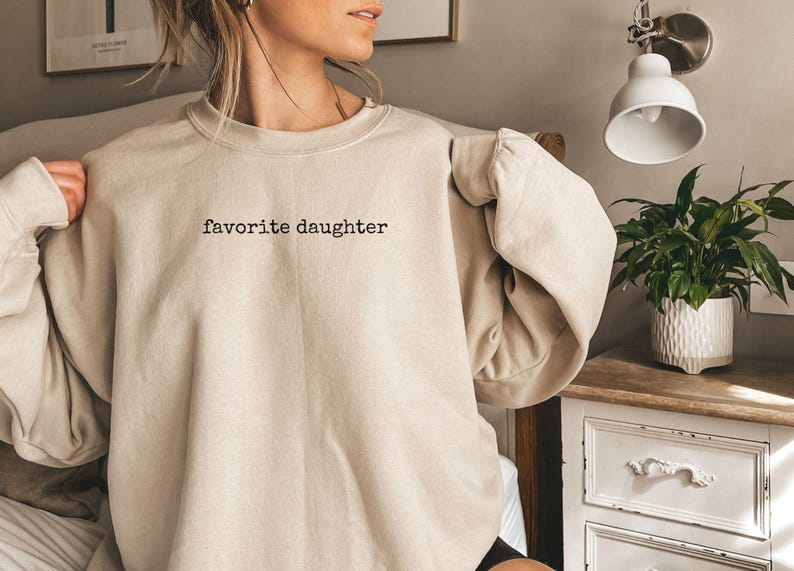 Może przedstawiać: Beżowa bluza z okrągłym dekoltem z czarnym napisem "favorite daughter". Bluza ma luźny kr&oacute;j i długie rękawy. Zdjęcie zrobione w pomieszczeniu.