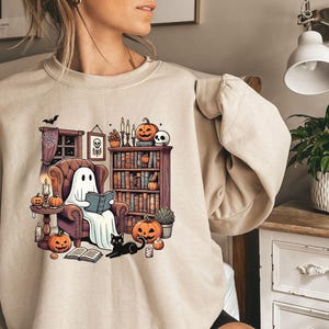 May include: Beige sweatshirt featuring a Halloween-themed graphic. The design shows a ghost reading in a cozy armchair surrounded by pumpkins, candles, and a black cat. A bookshelf and spooky decor complete the scene, perfect for fall.