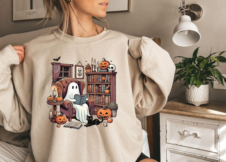 May include: Beige sweatshirt featuring a Halloween-themed graphic. The design shows a ghost reading in a cozy armchair surrounded by pumpkins, candles, and a black cat. A bookshelf and spooky decor complete the scene, perfect for fall.