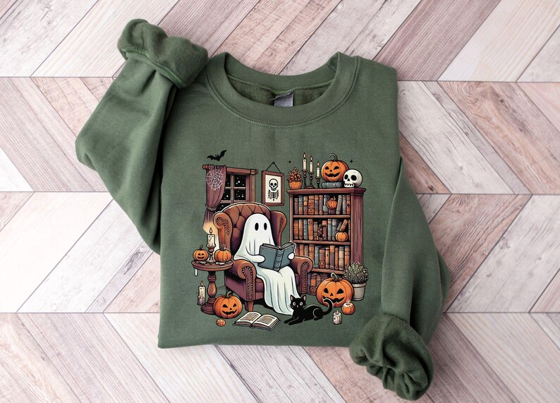 May include: Green sweatshirt featuring a cartoon ghost reading a book in a cozy, Halloween-themed room. The design includes pumpkins, candles, a bookshelf, and a black cat, creating a spooky and inviting atmosphere. Perfect for Halloween.