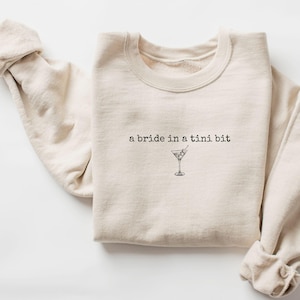 May include: A beige sweatshirt with the text "a bride in a tini bit" and a small illustration of a martini glass.