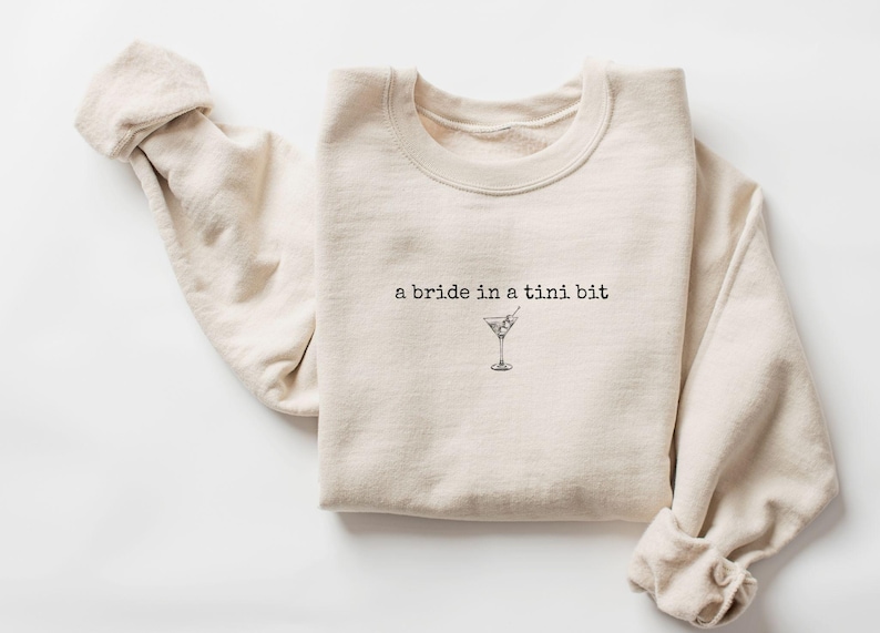 May include: A beige sweatshirt with the text "a bride in a tini bit" and a small illustration of a martini glass.