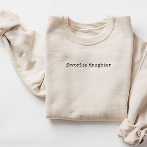 Może przedstawiać: Kremowa bluza z napisem "favorite daughter" wydrukowanym na czarno. Długie rękawy są podwinięte. Bluza jest starannie złożona na białej powierzchni.