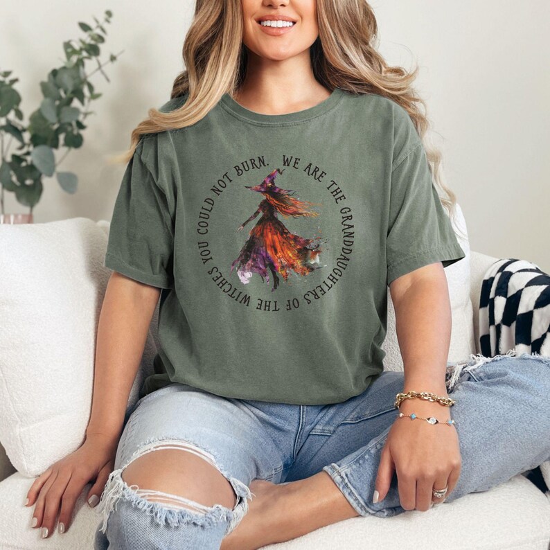 Pu&ograve; includere: Una donna indossa una maglietta verde con un'immagine di una strega e il testo "You could not burn. We are the granddaughters of the witches."