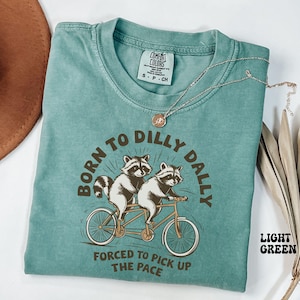 May include: Light green t-shirt featuring a graphic of two raccoons riding a tandem bicycle. The text "Born to Dilly Dally" is above the raccoons and "Forced to Pick Up the Pace" is below them.