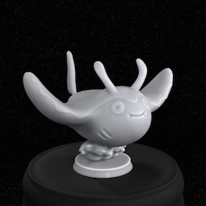 May include: A white 3D printed figurine of a cartoon stingray with a smiling face and two small horns on its head. The figurine is standing on a round base.