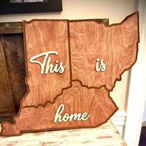May include: Wooden wall art in the shape of three states, with the words "This is home" in white script. The wood has a warm, reddish-brown tone, and the piece is displayed on a distressed white and brown cabinet.