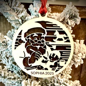 May include: A wooden Christmas ornament featuring a cut-out design of Santa Claus, a rocking horse, and other festive elements. The ornament is personalized with the name "SOPHIA" and the year "2025". It hangs from a red ribbon and is set against a snowy Christmas tree.