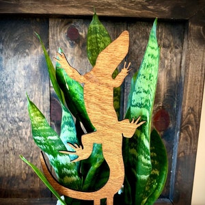 Puede incluir: Una estaca de planta de madera con forma de lagarto en una maceta blanca con una planta serpiente verde. El lagarto es marrón y tiene una cola larga.