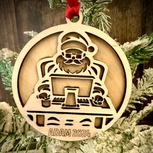 May include: A wooden Christmas ornament featuring a cartoon Santa Claus sitting at a desk with a laptop. The ornament is laser cut and has a personalized inscription that reads "ADAM 2024".