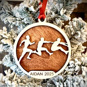 Peut inclure: Ornement de Noël rond en bois avec un motif découpé de trois athlètes en action. L'ornement a une bordure blanche et un centre en bois brun. Le nom "AIDAN 2025" est inscrit en bas. Un ruban rouge est attaché pour l'accrocher.