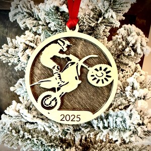 May include: A circular wooden Christmas decoration featuring a motocross rider. The design shows a cutout of a rider on a motorbike, with the year "2025" inscribed below. It is suspended by a red ribbon and set against a snowy evergreen background.