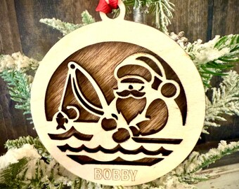 May include: A wooden Christmas bauble featuring a Santa Claus figure fishing with a rod and line. The bauble is brown with a white background and the name "BOBBY" is engraved on the bottom.