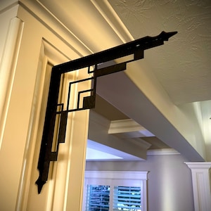 May include: A black metal decorative bracket with a geometric design. The bracket is mounted to a white wall.