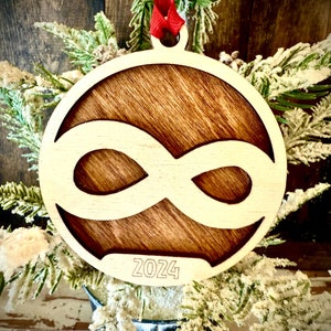 May include: A wooden Christmas ornament with a brown stain and a white infinity symbol cut out. The ornament is hanging from a red ribbon and has the year 2024 engraved on the bottom.