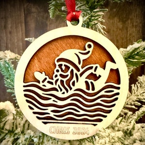 May include: A round wooden Christmas ornament with a laser-cut design of Santa Claus swimming in the ocean. The ornament is brown with a wood grain finish and has the text "CHRIS 2024" engraved on the bottom.