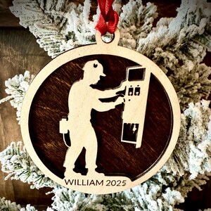 May include: A wooden Christmas ornament featuring a silhouette of a worker operating machinery. The ornament has a dark brown interior and a light brown exterior with the name "WILLIAM 2025" at the bottom. A red ribbon is attached for hanging.
