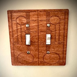 May include: A double light switch cover made of wood with an intricate geometric design. The cover is a dark reddish-brown color and has two white toggle switches.