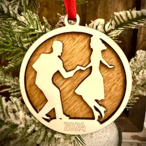 May include: Wooden Christmas ornament featuring a dancing couple silhouette inside a circular frame. The ornament has a natural wood grain background and a light-colored border. The year "2024" is engraved at the bottom. A red ribbon is attached for hanging.