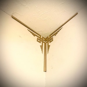 Puede incluir: Una moldura de esquina decorativa de color dorado con un diseño geométrico. La moldura está unida a la esquina de un techo y una pared blancos.