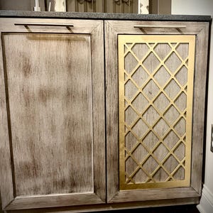 May include: A two-door cabinet with a distressed wood finish. One door features a gold diamond lattice design. The cabinet has a dark countertop and silver handles. The overall aesthetic is rustic and elegant.