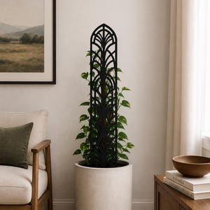 May include: A black metal plant trellis with an arched top, featuring a decorative leaf design. The trellis supports a climbing plant with green leaves, placed in a large, light-coloured cylindrical pot. The scene is set indoors.
