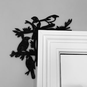 Può includere: Decorazione da parete in metallo nero a forma di silhouette di uccello con un motivo a ramo. Il decoro è montato su uno stipite di porta bianco.
