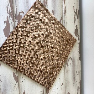 May include: A square piece of wood with a geometric pattern burned into the surface. The pattern is made up of small squares and rectangles, creating a repeating design.