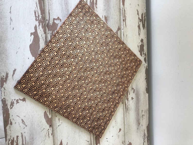 May include: A square piece of wood with a geometric pattern burned into the surface. The pattern is made up of small squares and rectangles, creating a repeating design.