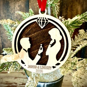 May include: A round wooden ornament with a silhouette of a couple getting engaged. The ornament is decorated with a diamond and the text "Jarod & Lindsey 2024".