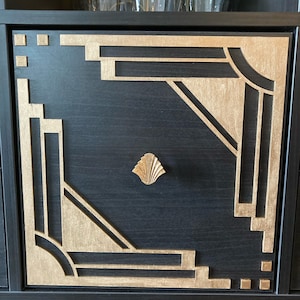 May include: A black cabinet door with a gold geometric design. The door has a decorative gold knob in the center.