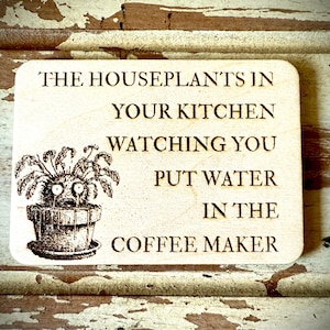 May include: A wooden sign with a black and white illustration of a houseplant with eyes. The sign reads "The houseplants in your kitchen watching you put water in the coffee maker."
