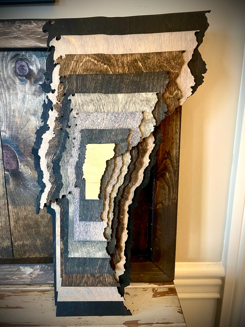 Puede incluir: Una pieza de arte de pared de madera en capas con forma del estado de Vermont. La madera est&aacute; te&ntilde;ida en varios tonos de marr&oacute;n, negro y gris.