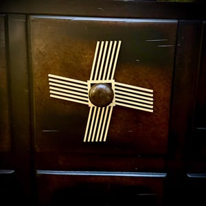 May include: Close-up of a dark brown wooden cabinet door with a decorative cross-shaped design. The cross is made of cream-colored stripes and a central dark brown knob. The wood grain is visible.