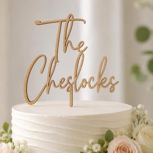 May include: A light brown wooden cake topper featuring the text The Cheslocks in a cursive script font. The topper is positioned on top of a white frosted cake with horizontal texture, decorated with white and light pink roses and greenery.