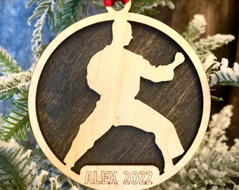 May include: A round wooden Christmas bauble featuring a silhouette of a person in a karate pose. The bauble is decorated with a dark wood stain and has the text "ALEX 2022" engraved on the bottom.
