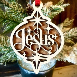 May include: A wooden Christmas ornament shaped like a bauble with the word "Jesus" cut out in a decorative font. The ornament is decorated with stars and has a red ribbon attached.