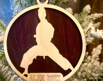 May include: A wooden Christmas bauble featuring a silhouette of a person in a karate pose. The bauble is painted in shades of brown and red and has the name "ALEX 2022" engraved on the bottom.