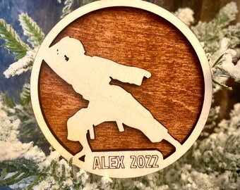 May include: A wooden Christmas bauble featuring a silhouette of a person in a karate pose. The bauble is brown with a white silhouette and the text "ALEX 2022" is engraved on the bottom.