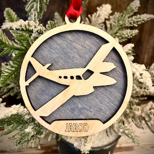 May include: A wooden Christmas ornament with a silhouette of a small airplane and the name "JAROD" inscribed on the bottom. The ornament is hanging from a red ribbon on a snowy pine branch.
