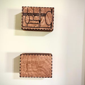 May include: Two wooden wall-mounted boxes with geometric designs. The top box features a design with circles and lines, while the bottom box has a different pattern. Both boxes are made of light brown wood with dark brown etched details.