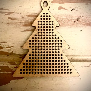 May include: A wooden Christmas tree ornament with a hole at the top for hanging. The ornament is made of light brown wood and has a grid of small holes throughout the surface.