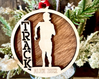 Peut inclure: Une décoration de Noël en bois avec une silhouette de coureur et le mot "TRACK" sur le côté gauche. La décoration est personnalisée avec le nom "ALEX" et l'année "2022".