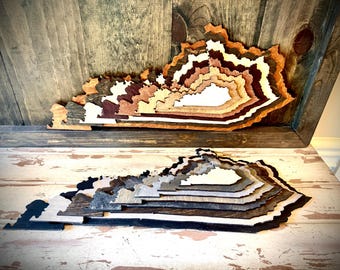 Può includere: Due mappe in legno a strati del Kentucky, una nei toni del marrone e del bianco, l'altra in grigio e nero. Le mappe sono esposte su una superficie di legno rustico, che mostra il contorno dello stato.