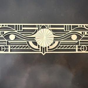 May include: A rectangular light-colored wooden wall decoration featuring intricate geometric cutouts and lines. A circular sunburst-patterned wooden piece is mounted in the center, surrounded by symmetrical patterns of horizontal bars, curves, and circles.