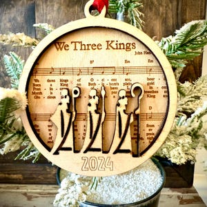 May include: A wooden Christmas bauble featuring a cut-out design of the three wise men, the words "We Three Kings" and the year "2024". The bauble is decorated with sheet music from the carol "We Three Kings".