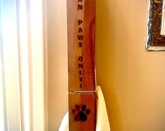 以下が含まれることがあります： 「CLEAN PAWS ONLY！」の文字と足跡のデザインが施された木製のタオルホルダー。ホルダーには、濃い茶色の足跡がプリントされた白いタオルが取り付けられています。ホルダーは壁に取り付けられています。